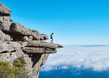 A lone person on a dramatic cliff edge gazing over a sea of clouds under a bright blue sky.