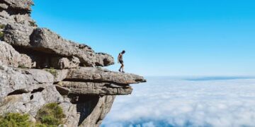 A lone person on a dramatic cliff edge gazing over a sea of clouds under a bright blue sky.
