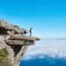 A lone person on a dramatic cliff edge gazing over a sea of clouds under a bright blue sky.