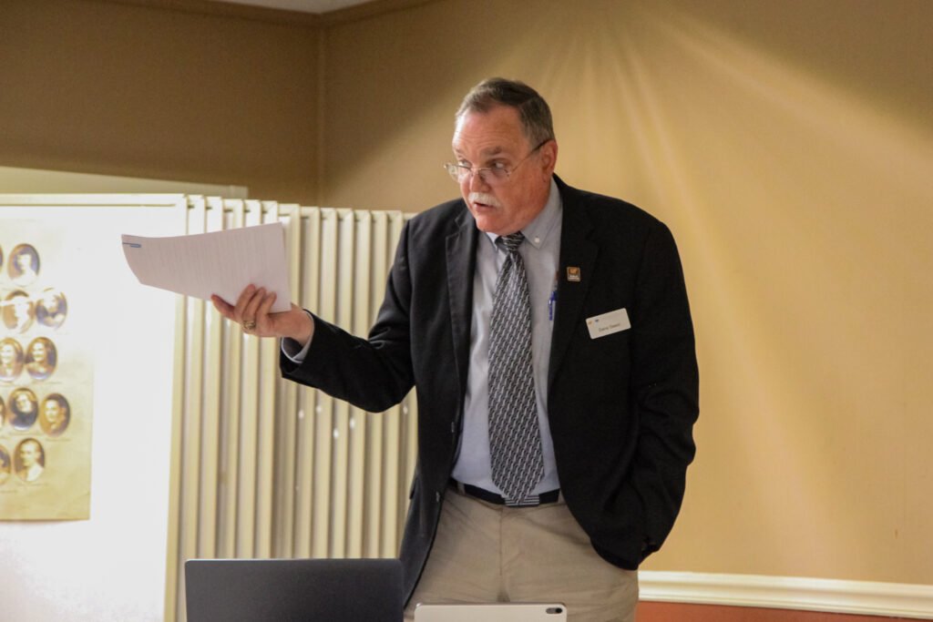 Dana Deem stands at the front of the room in a suit and tie, holding documents while addressing the Atwood board.