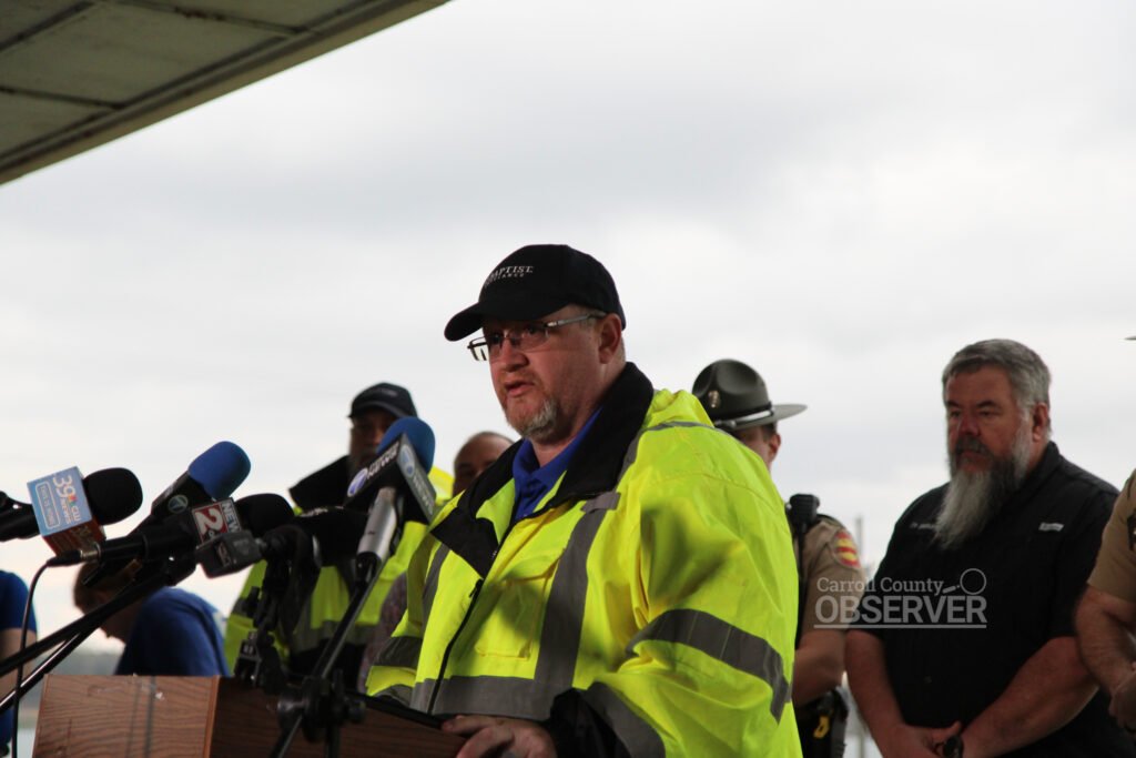 A man in a yellow high-visibility jacket speaks at a podium during a press briefing about a fatal school bus crash in Carroll County, Tennessee.