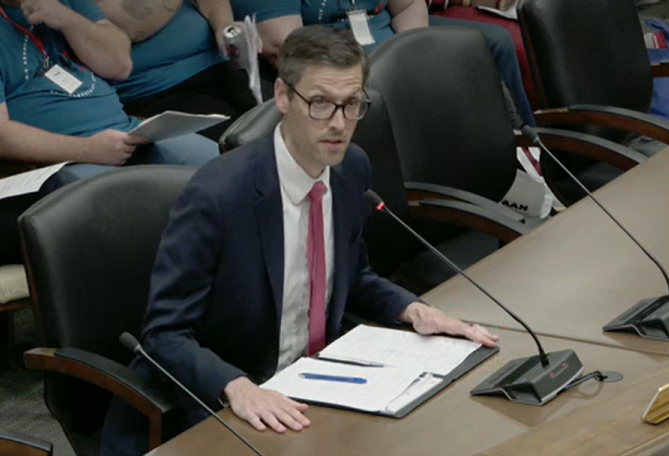 A man in a navy suit and red tie speaks at a committee table with documents in front of him.