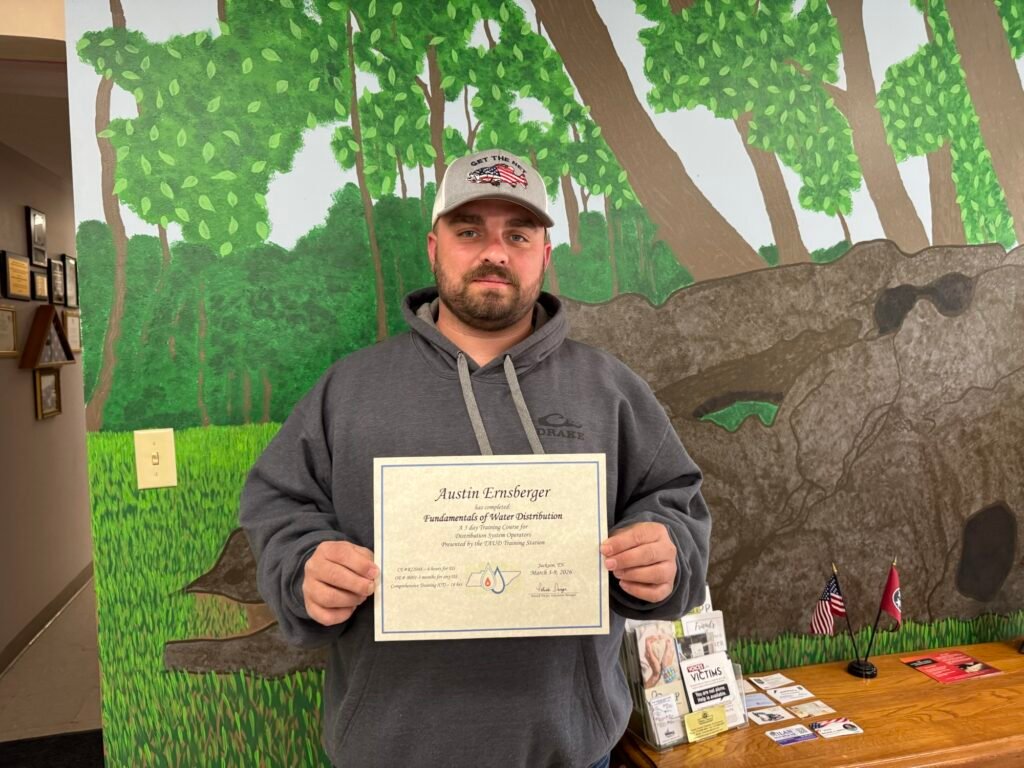 Austin Ernsberger holds his Fundamentals of Water Distribution certificate in front of a mural at Hollow Rock Town Hall.