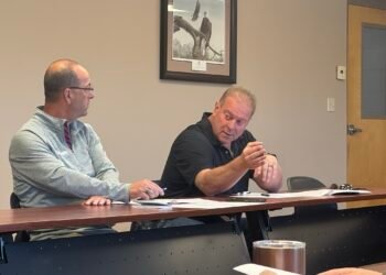 Two men seated at a conference table during a county committee meeting, with the man on the right gesturing while speaking.