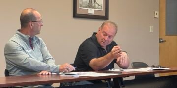 Two men seated at a conference table during a county committee meeting, with the man on the right gesturing while speaking.