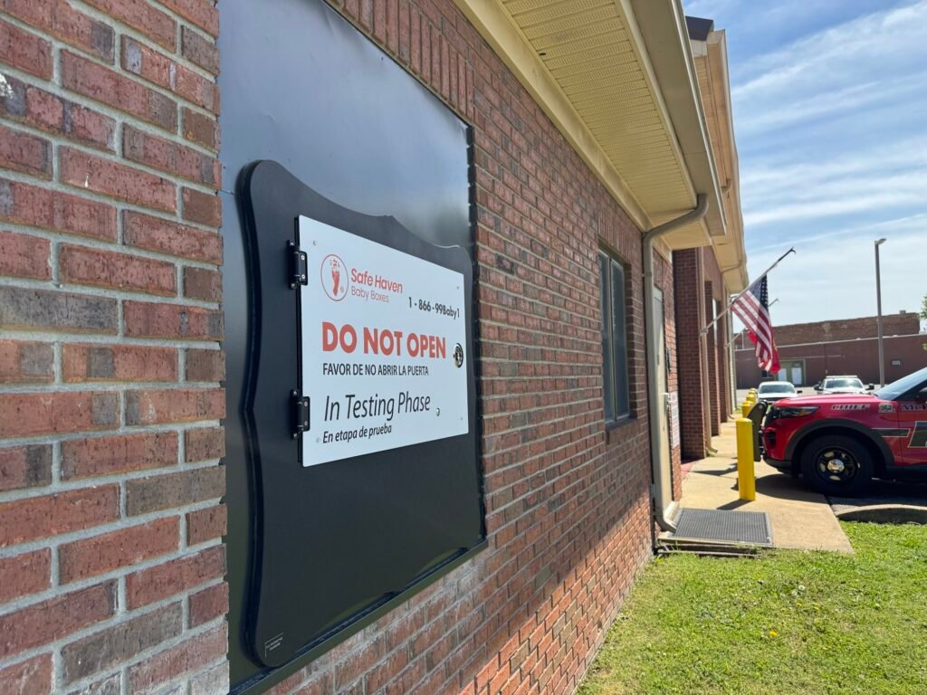 Close-up of a Safe Haven baby box door on a brick wall with a sign reading "Do Not Open, In Testing Phase" in English and Spanish.