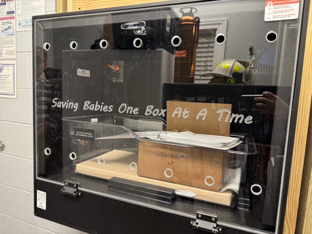 Interior of a Safe Haven baby box with a bassinet labeled "In Loving Memory of Cameron C. Scott" and a Tennessee kit inside.