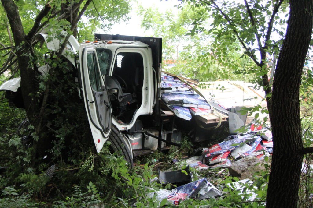 Crushed cab of a Freightliner flatbed truck rests against a tree after crashing off State Route 22.